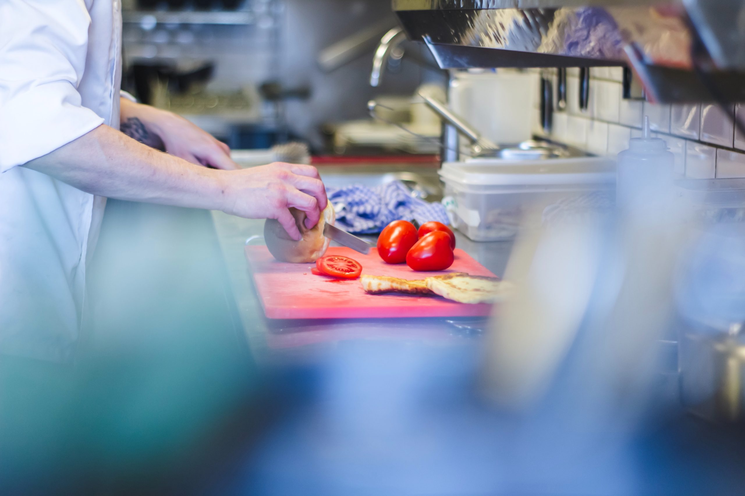 Chef wearing shoe covers in a restaurant kitchen slicing bread with a knife on a cutting board with tomatoes.