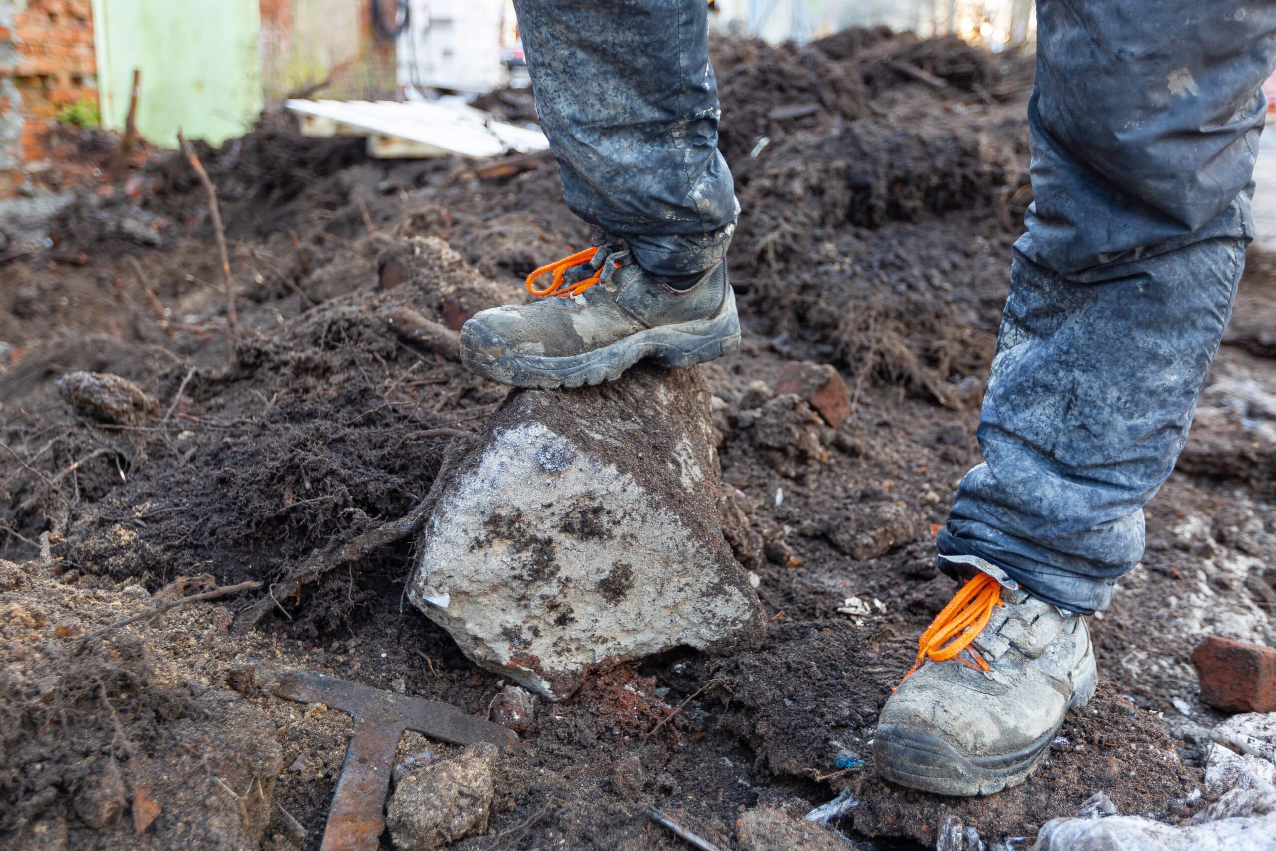Worker in dirty uniform and shoes with orange laces is standing on a stone in a construction site, showing the need for shoe covers before going indoors.