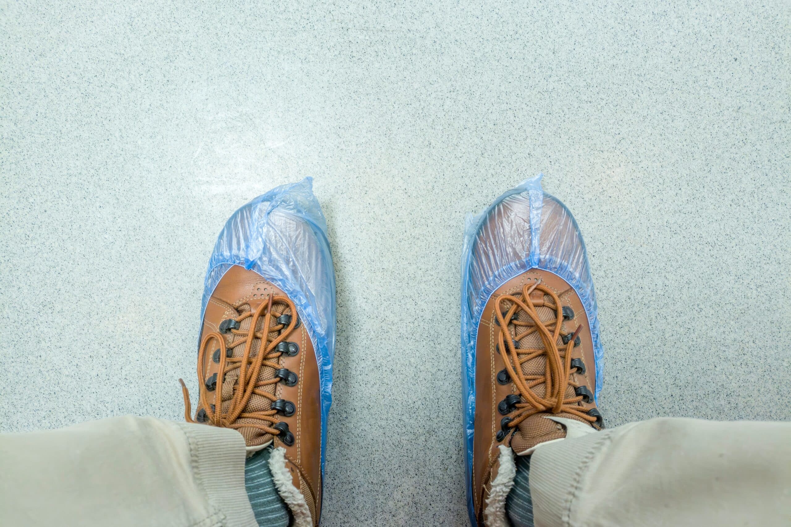 Feet in brown shoes with blue disposable shoe covers on a clean floor, illustrating hygiene and safety in public or medical spaces.